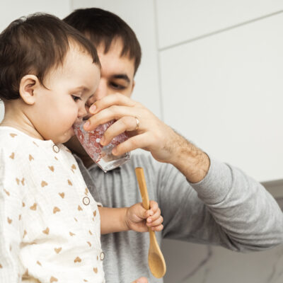 A caring father stands in the kitchen with a daughter in his arms, singing his baby with water from a glass.